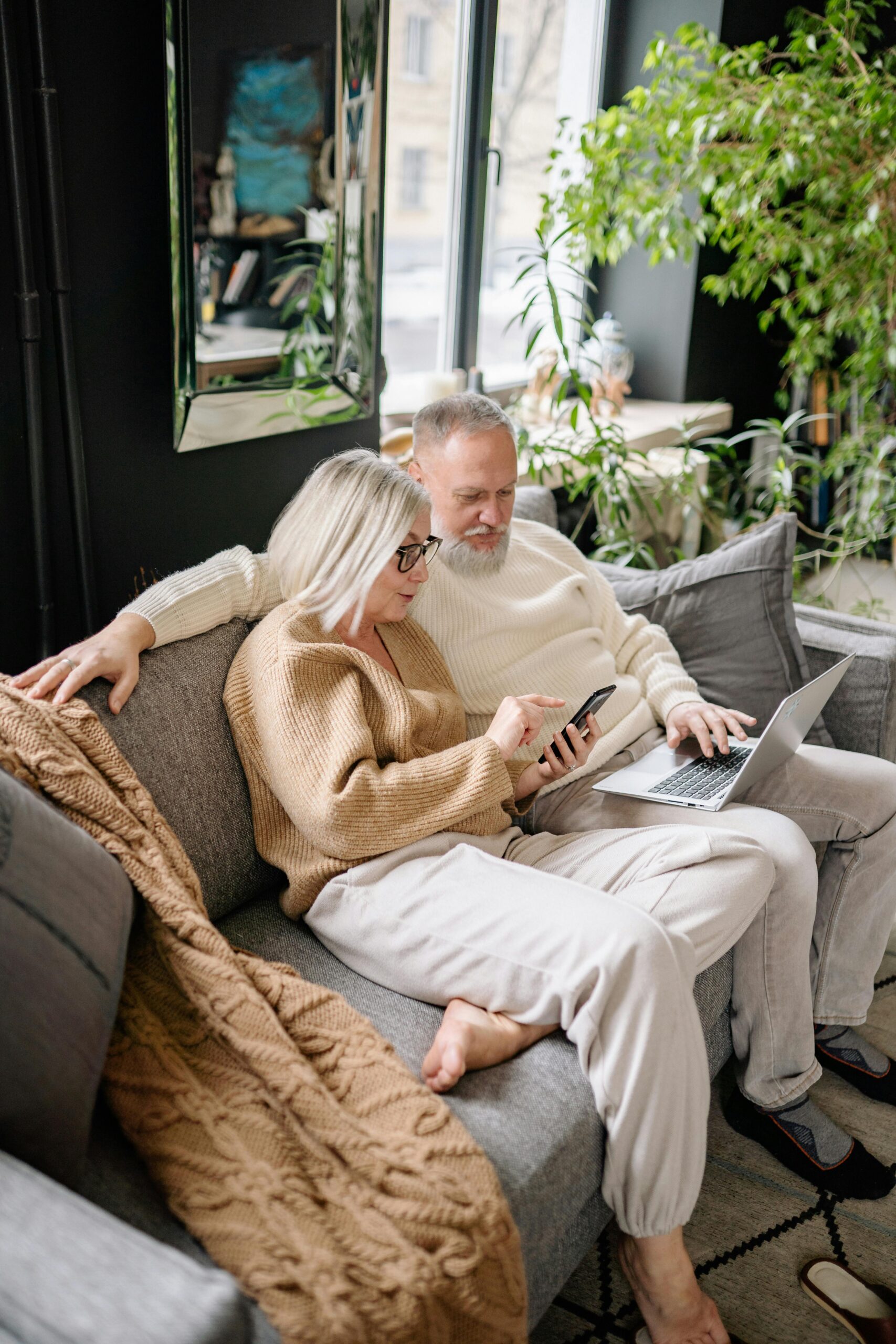 Senior couple sitting on couch using laptop and smartphone together in cozy home setting.